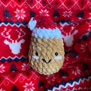May include: A brown crocheted plush toy shaped like a pineapple wearing a red and white crocheted Santa hat. The toy has a smiling face with black eyes and white cheeks. The toy is sitting on a red and white patterned blanket.