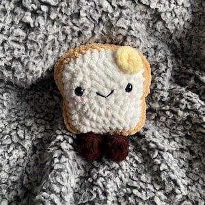 May include: A crocheted slice of toast with a smiling face, rosy cheeks, and a pat of yellow butter. The toast has brown crusts and brown feet. The plush toy is set against a grey, textured background.