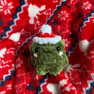 May include: A handmade green crochet frog wearing a red and white Santa hat. The frog has black eyes and pink cheeks. The frog is on a red and white patterned blanket.