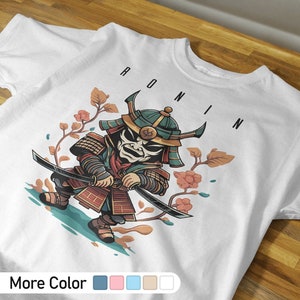 Może przedstawiać: Biała koszulka z kreskówkową ilustracją samuraja trzymającego miecz. Samuraj ma na sobie zielony hełm i czerwoną zbroję. Tekst "RONIN" jest nadrukowany czarnym kolorem nad ilustracją.