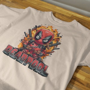 Puede incluir: Una camiseta beige con una imagen de dibujos animados de Deadpool con un traje rojo y negro, rodeado de llamas y espadas. El texto "DEADPOOL" est&aacute; impreso en blanco y negro debajo de la imagen.