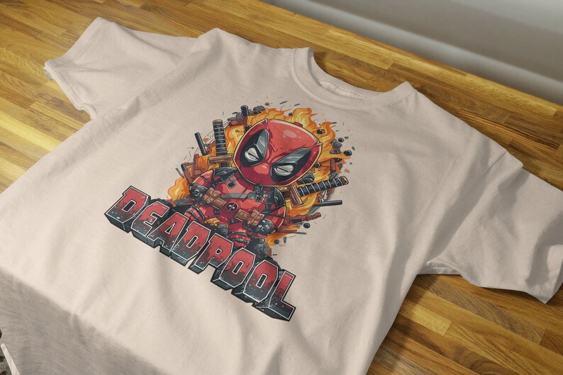 Puede incluir: Una camiseta beige con una imagen de dibujos animados de Deadpool con un traje rojo y negro, rodeado de llamas y espadas. El texto "DEADPOOL" est&aacute; impreso en blanco y negro debajo de la imagen.