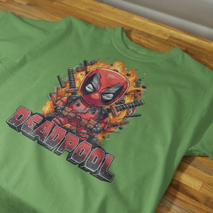 Puede incluir: Camiseta verde con una imagen de dibujos animados de Deadpool con un traje rojo y negro, rodeado de llamas y armas. El texto "DEADPOOL" est&aacute; impreso en blanco y negro debajo de la imagen.