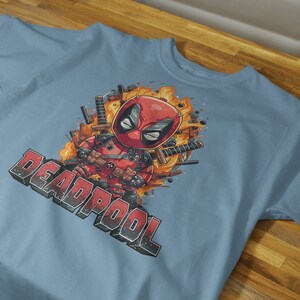 Puede incluir: Una camiseta azul claro con un estampado gr&aacute;fico de Deadpool con un traje rojo y negro, rodeado de llamas y espadas. El texto "DEADPOOL" est&aacute; impreso en negro debajo del gr&aacute;fico.