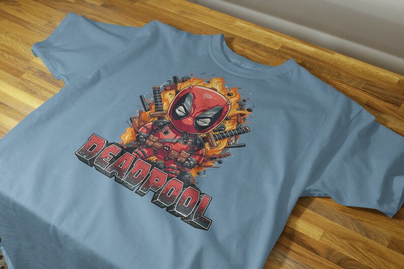Puede incluir: Una camiseta azul claro con un estampado gr&aacute;fico de Deadpool con un traje rojo y negro, rodeado de llamas y espadas. El texto "DEADPOOL" est&aacute; impreso en negro debajo del gr&aacute;fico.
