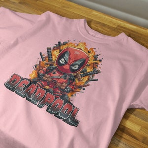 Puede incluir: Camiseta rosa con un estampado gr&aacute;fico de Deadpool en una explosi&oacute;n de fuego. El texto "DEADPOOL" est&aacute; impreso debajo de la imagen.