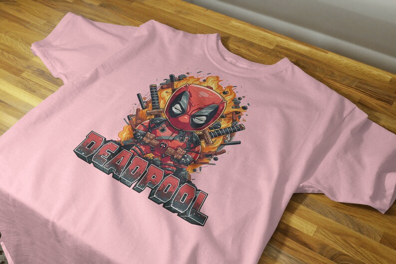 Puede incluir: Camiseta rosa con un estampado gr&aacute;fico de Deadpool en una explosi&oacute;n de fuego. El texto "DEADPOOL" est&aacute; impreso debajo de la imagen.