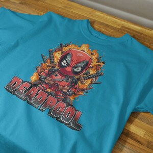 Puede incluir: Una camiseta azul turquesa con un estampado gr&aacute;fico de Deadpool, un personaje de superh&eacute;roe, en estilo de dibujos animados. El gr&aacute;fico presenta a Deadpool con espadas y llamas. El texto "DEADPOOL" est&aacute; impreso en blanco y negro debajo del gr&aacute;fico.
