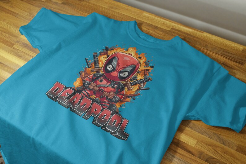Puede incluir: Una camiseta azul turquesa con un estampado gr&aacute;fico de Deadpool, un personaje de superh&eacute;roe, en estilo de dibujos animados. El gr&aacute;fico presenta a Deadpool con espadas y llamas. El texto "DEADPOOL" est&aacute; impreso en blanco y negro debajo del gr&aacute;fico.