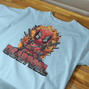 Puede incluir: Camiseta azul claro con un estampado gr&aacute;fico de Deadpool con un traje rojo y negro, rodeado de llamas y espadas. El texto "DEADPOOL" est&aacute; impreso en negro debajo del gr&aacute;fico.
