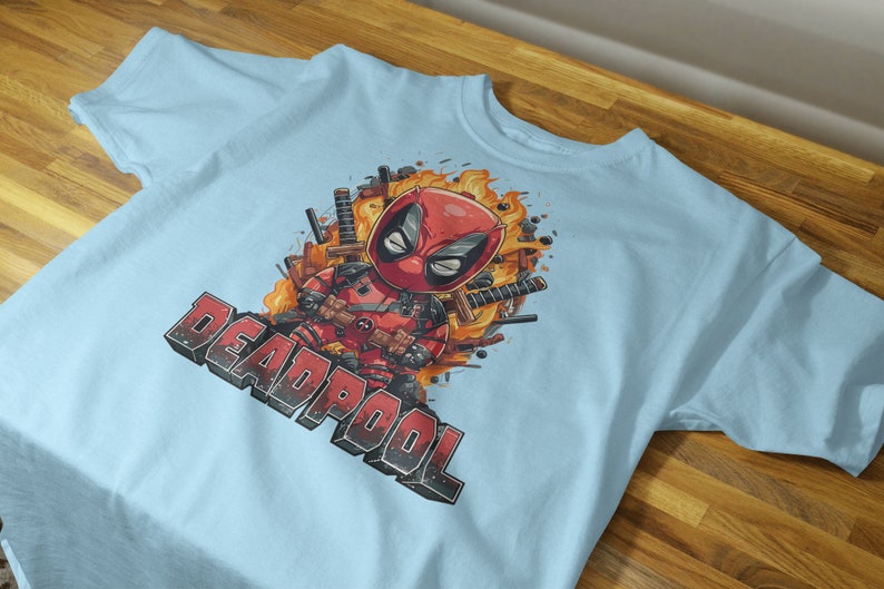 Puede incluir: Camiseta azul claro con un estampado gr&aacute;fico de Deadpool con un traje rojo y negro, rodeado de llamas y espadas. El texto "DEADPOOL" est&aacute; impreso en negro debajo del gr&aacute;fico.