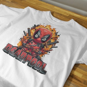 Puede incluir: Camiseta blanca con una imagen de dibujos animados de Deadpool con un traje rojo y negro, rodeado de llamas y espadas. El texto "DEADPOOL" est&aacute; impreso en negro debajo de la imagen.