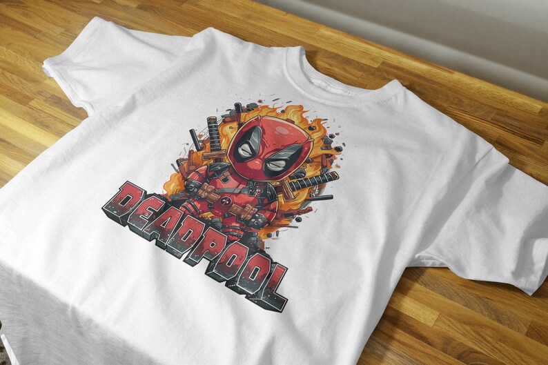 Puede incluir: Camiseta blanca con una imagen de dibujos animados de Deadpool con un traje rojo y negro, rodeado de llamas y espadas. El texto "DEADPOOL" est&aacute; impreso en negro debajo de la imagen.