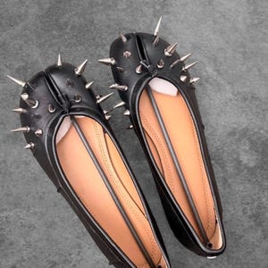 Puede incluir: Un par de bailarinas negras con punta dividida que presentan un adorno de púas metálicas de tono plateado en la parte delantera. El interior de los zapatos está forrado con un material color canela y cada uno incluye un inserto estructural negro.