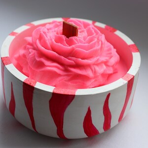 May include: A pink candle in a white and red striped bowl. The candle is shaped like a rose and has a wooden wick. The bowl is round and has a painted design.