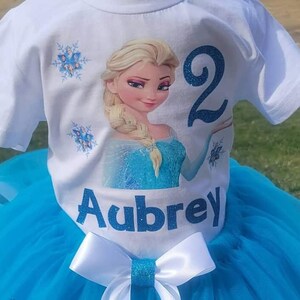 May include: White t-shirt with a graphic of a blonde princess in a blue dress, the number 2, and the name "Aubrey" in blue glitter. The shirt is paired with a blue tutu skirt and a white bow with a glitter center.