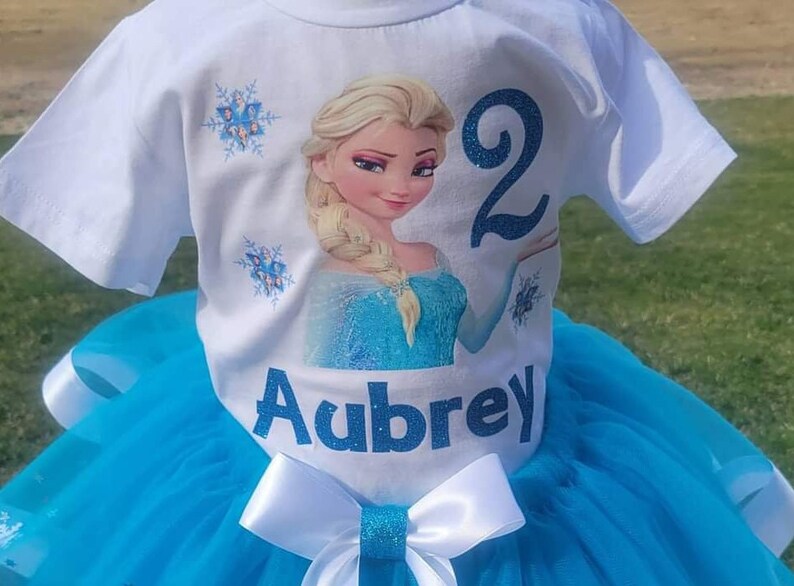 May include: White t-shirt with a graphic of a blonde princess in a blue dress, the number 2, and the name "Aubrey" in blue glitter. The shirt is paired with a blue tutu skirt and a white bow with a glitter center.