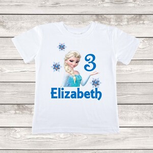 May include: White t-shirt featuring a graphic of a princess with blonde hair and a blue dress. The number "3" and the name "Elizabeth" are printed in blue. Decorative snowflakes are also included in the design.