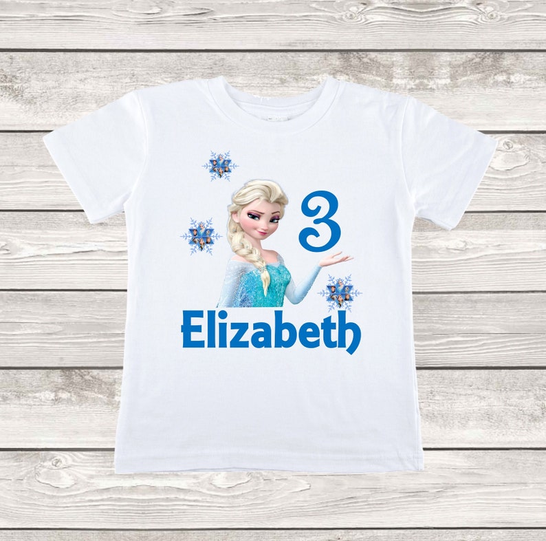 May include: White t-shirt featuring a graphic of a princess with blonde hair and a blue dress. The number "3" and the name "Elizabeth" are printed in blue. Decorative snowflakes are also included in the design.