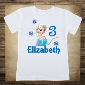 May include: White t-shirt featuring a graphic of a blonde princess in a blue dress, with the name "Elizabeth" and the number "3" in blue. The design includes snowflakes.