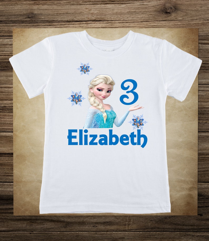 May include: White t-shirt featuring a graphic of a blonde princess in a blue dress, with the name "Elizabeth" and the number "3" in blue. The design includes snowflakes.
