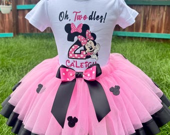 May include: A white short-sleeved bodysuit featuring the text Oh, Toodles! and the number 2 with a Minnie Mouse graphic and the name Caleigh. The bodysuit is paired with a pink tulle tutu skirt featuring black ribbon trim and black mouse ears.