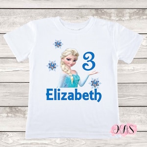 May include: White t-shirt featuring a graphic of a blonde princess in a blue dress, the number 3, and the name "Elizabeth" in blue. The design includes blue snowflake accents. A children's birthday shirt.