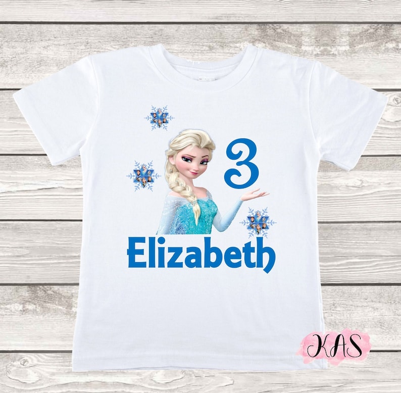 May include: White t-shirt featuring a graphic of a blonde princess in a blue dress, the number 3, and the name "Elizabeth" in blue. The design includes blue snowflake accents. A children's birthday shirt.
