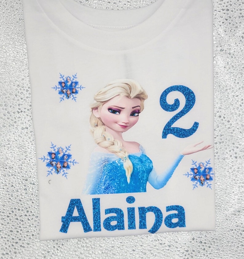May include: White t-shirt featuring a graphic of a blonde woman in a blue dress, with the number "2" and the name "Alaina" in blue glitter. The shirt also has snowflake designs.