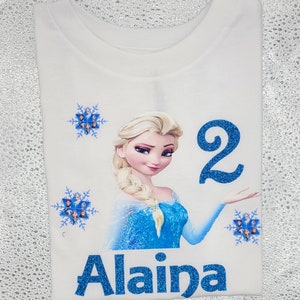 May include: White t-shirt featuring a glittery blue graphic of Elsa from Frozen, the number 2, and the name Alaina. The design includes blue snowflakes with character images. The shirt is a children's birthday theme.
