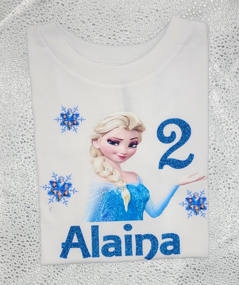 May include: White t-shirt featuring a glittery blue graphic of Elsa from Frozen, the number 2, and the name Alaina. The design includes blue snowflakes with character images. The shirt is a children's birthday theme.