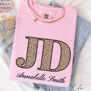 May include: A light pink t-shirt with the initials "JD" in a leopard print design and the name "Annabelle Smith" in black script. The shirt is folded on top of blue jeans, with a gold necklace and the text "Color: Blossom".