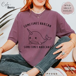 May include: A berry-colored short-sleeve t-shirt featuring a black line-art illustration of a narwhal in the center. The text "SOMETIMES NARCAN SOMETIMES NARCANT" is printed in black above and below the narwhal graphic on the front of the shirt.