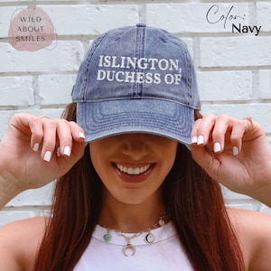 May include: A navy blue baseball cap with a distressed finish featuring the white embroidered text "ISLINGTON, DUCHESS OF" on the front panel. The cap has a curved brim and a structured crown, showcasing a classic six-panel design with top stitching.