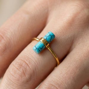Könnte beinhalten: Ein goldfarbener Ring mit einem rechteckigen, türkisfarbenen Stein mit dunkelblauen Adern. Der Stein wird von einem goldenen Band gehalten. Der Ring ist ein Schmuckstück.