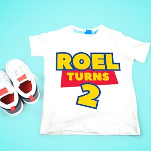 May include: White t-shirt with a red, yellow, and blue graphic that says "Roel Turns 2". A pair of white sneakers with red and blue accents are on a light blue background.