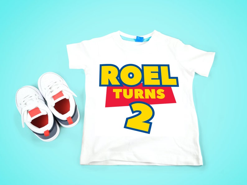 May include: White t-shirt with a red, yellow, and blue graphic that says "Roel Turns 2". A pair of white sneakers with red and blue accents are on a light blue background.