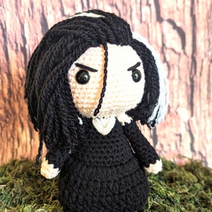 May include: Crocheted doll with black hair and a black dress. The doll has a stern expression with black eyes and a white face. The hair is styled with a white streak. The doll is sitting on a bed of green moss.