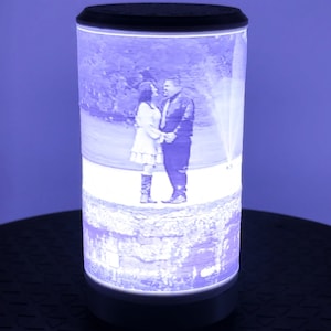 May include: A cylindrical lamp with a black top and base, displaying a black and white image of a couple. The lamp emits a soft, cool-toned light, illuminating the image and the surrounding area. The lamp is on a dark surface.