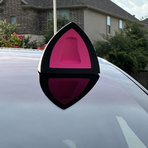 Könnte beinhalten: Ein schwarzer und rosa Autoantennenaufsatz in Form von Katzenohren. Die Ohren haben eine schwarze Umrandung mit einem rosa Innenraum und einer helleren rosa Mitte. Der Aufsatz ist auf einem weißen Auto montiert.
