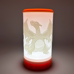 Puede incluir: Lámpara cilíndrica con base y parte superior naranja, y cuerpo blanco translúcido. El cuerpo presenta una ilustración detallada y clara de un Charizard. La lámpara está iluminada desde dentro, proyectando un brillo cálido.