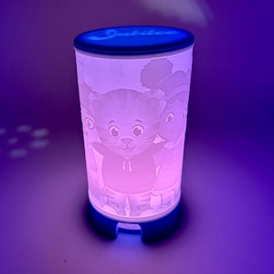 May include: A white night light with a blue base and a blue top. The night light has a design of the characters from the show "The Tiger Who Came to Tea".