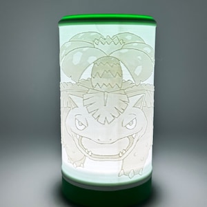 Könnte beinhalten: Zylindrische Lampe mit grünem Sockel und Zierleisten, mit einem geätzten Design eines Zeichentrickcharakters mit einer Blume und einem lächelnden Gesicht. Die Lampe strahlt ein sanftes, weißes Leuchten aus. Das Design ist detailliert und die Lampe ist grün.