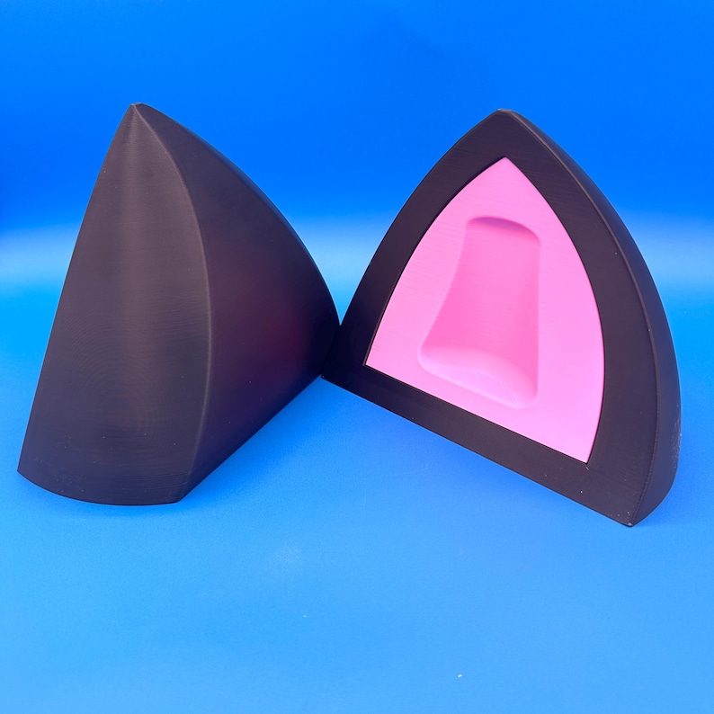 K&ouml;nnte beinhalten: Zwei schwarze Katzenohren-Accessoires, eines massiv und das andere aufgeschnitten, um ein rosa Innenleben zu zeigen. Die Ohren heben sich vor einem leuchtend blauen Hintergrund ab und erzeugen einen auff&auml;lligen Kontrast. Das Design ist schlicht und modern.