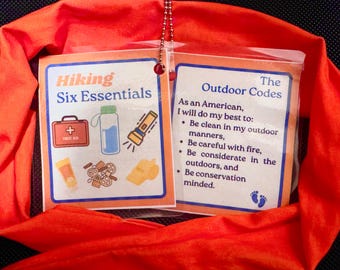 May include: A bright orange fabric pouch with two laminated cards attached. The left card has the title "Hiking Six Essentials" and shows illustrations of a first aid kit, a water bottle, and a flashlight. The right card has the title "The Outdoor Codes" and lists guidelines for outdoor etiquette, including being clean, careful with fire, and considerate of the outdoors.