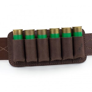 Può includere: Un porta cartucce in pelle marrone per fucile a canna liscia con sei cartucce verdi e in ottone. Il porta cartucce è attaccato a una cintura in pelle marrone. Le cuciture sono visibili sulla cintura e sul porta cartucce.