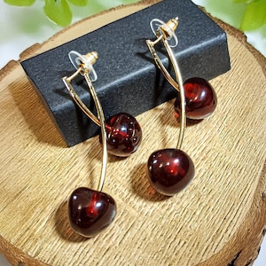 Puede incluir: Un par de pendientes de cereza. Cada pendiente presenta un tallo dorado con una cereza roja oscura y brillante en el extremo. Los pendientes se exhiben en una caja negra, sobre un fondo de madera con follaje verde.