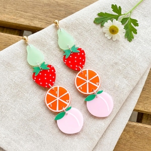Puede incluir: Un par de pendientes colgantes con temática de frutas. Los pendientes tienen postes dorados y están compuestos por formas acrílicas en capas. Las formas incluyen formas de pera verde claro, fresas rojas, rodajas de naranja y melocotones rosas.
