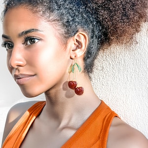 Puede incluir: Una mujer con cabello castaño rizado lleva una blusa naranja brillante sin mangas y pendientes de cereza colgantes. Los pendientes son de oro con dos cerezas rojas y hojas verdes.
