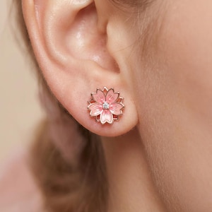 May include: A pair of pink flower stud earrings. Each earring features a pink enamel flower design with a small, clear gem in the center, set in a rose gold-colored metal. The earrings are shown on an ear.